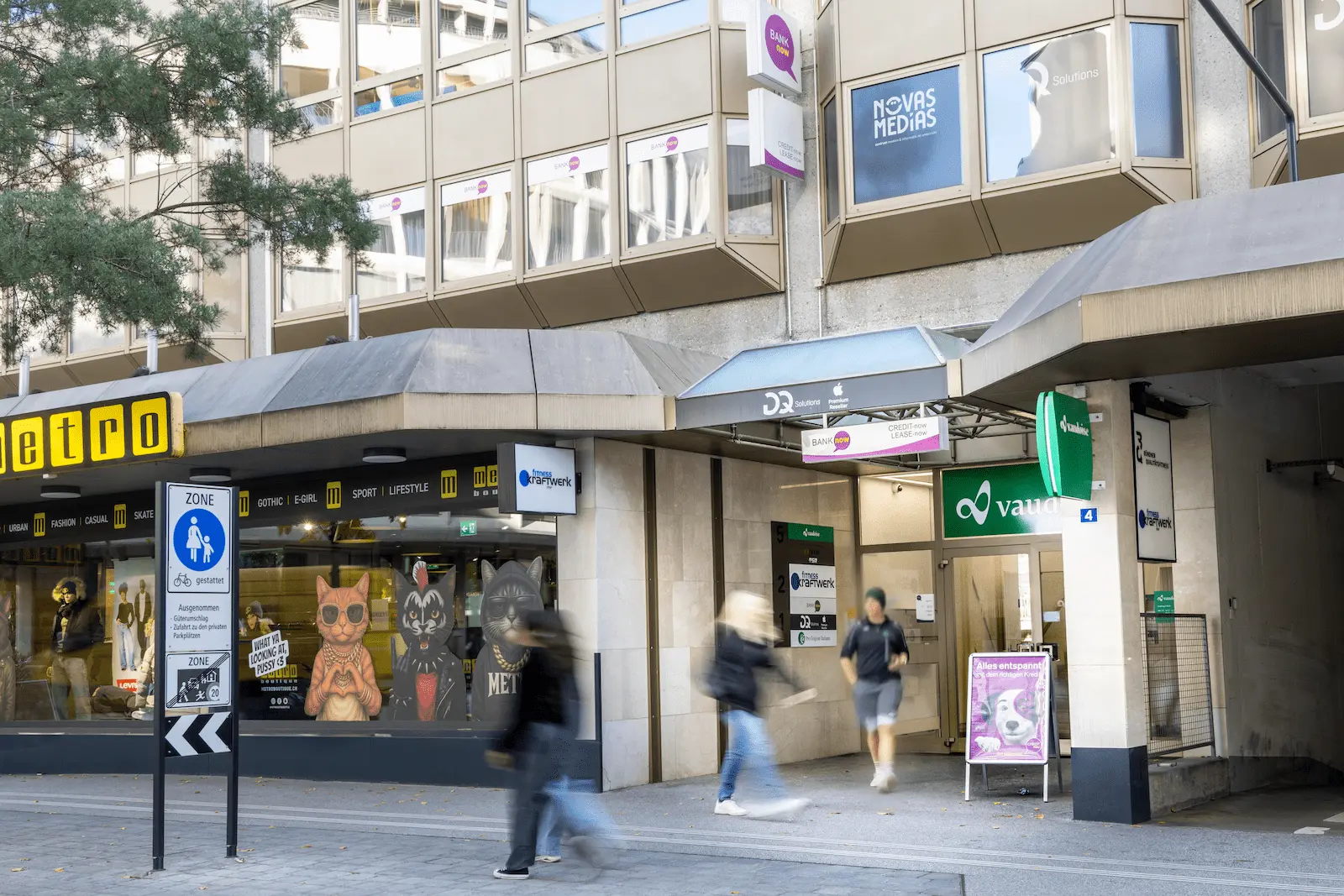 Breiter Blick auf den Eingang der BANK-now Filiale Chur mit Gebäudefassade und umliegender Strasse.