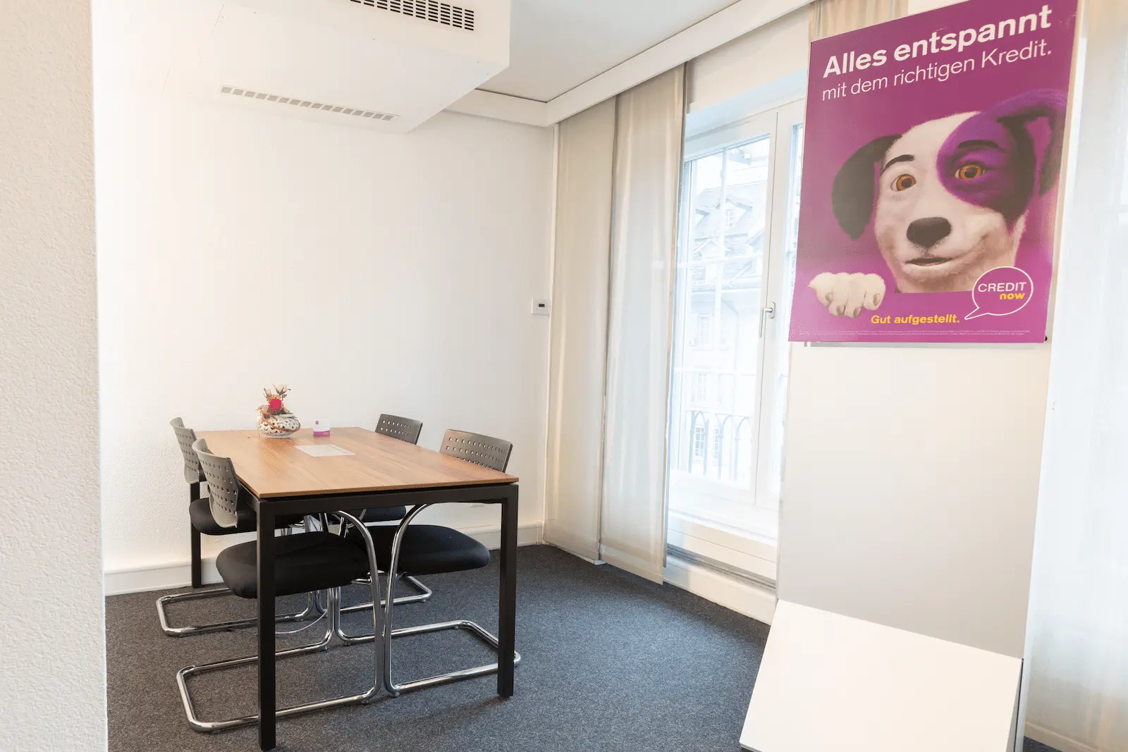 Besprechungsraum der BANK-now Filiale Bern mit Tisch, vier Stühlen und Poster an der Wand.